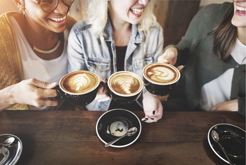 Three smiling women sitting at a coffeehouse holding their latte mugs up in the air