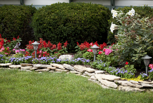 Landscaping in front of a home with colorful flowers in the front and large round bushes in the back against the house. The landscaping area is surrounded by a short wall of flat edging rock slabs.