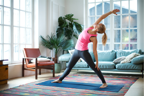 A woman in the middle of a yoga pose standing on an exercise mat in the middle of her modern living room.